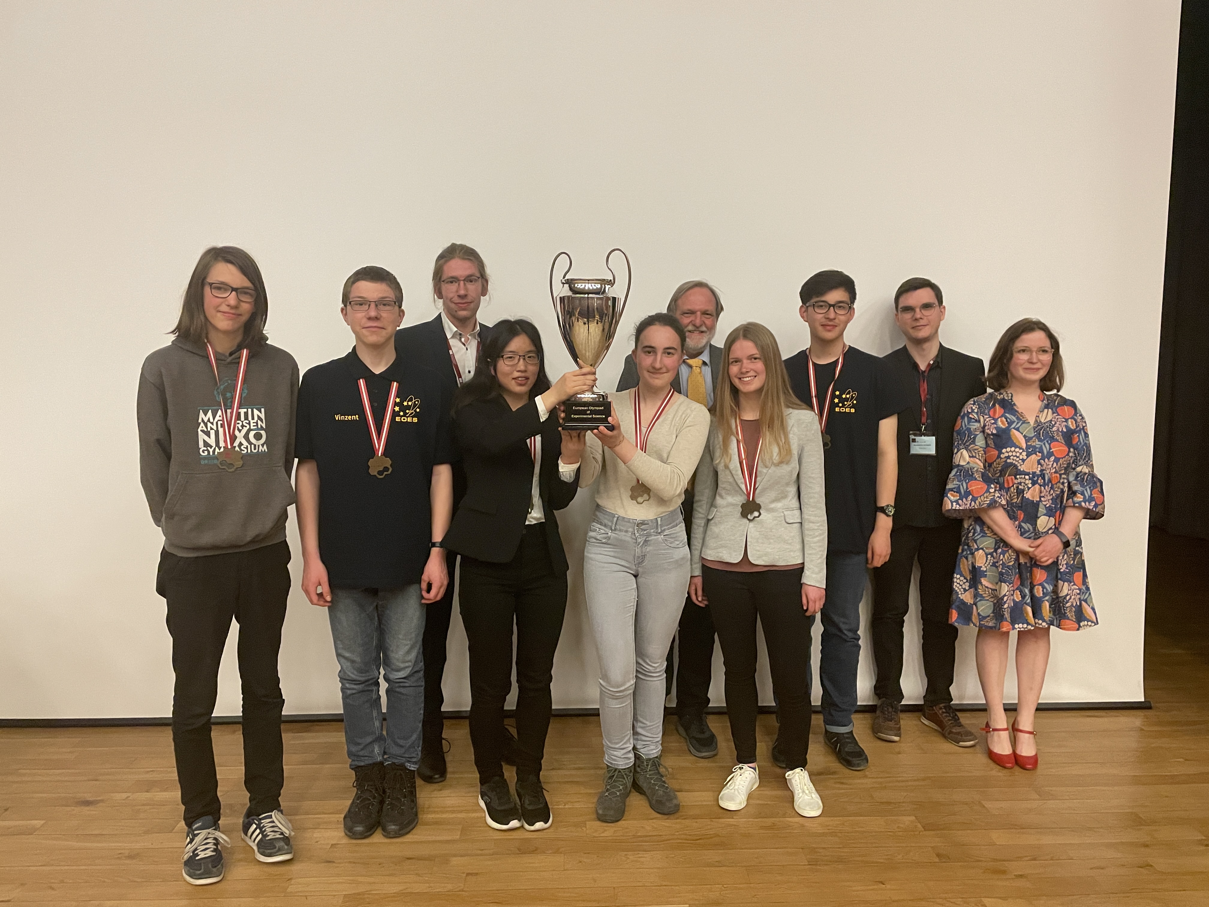 Die Jugendlichen der beiden deutschen Teams stehen zusammen mit ihren Mentoren lächelnd auf der Bühne. Sie tragen Medaillen um den Hals und halten einen Pokal hoch.   