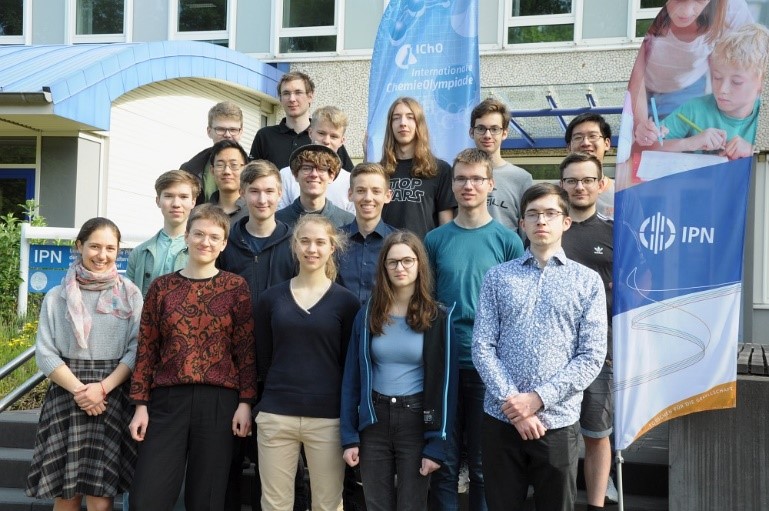 Die teilnehmenden Schülerinnen und Schüler der letzten Auswahlrunde zur 55. Internationalen ChemieOlympiade 2023 stehen vor dem Haupteingang des IPN. Rechts neben der Gruppe steht eine Fahne des IPN, die eine Frau zeigt, die einem Kind beim Lösen von einen Aufgabenblatt hilft. Hinter der Gruppe steht eine Fahne der Internationalen ChemieOlympiade.
