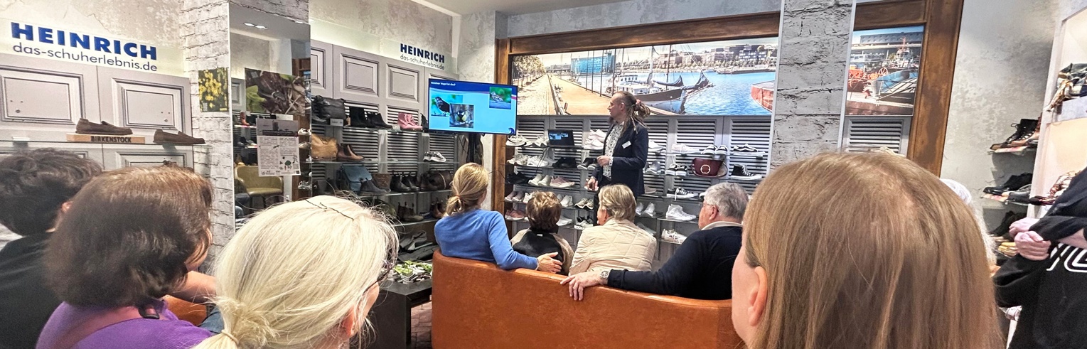 Dr. Michael Baum präsentiert an einem Monitor seinen Kurzvortrag vor Publikum im Schuhgeschäft Heinrich.